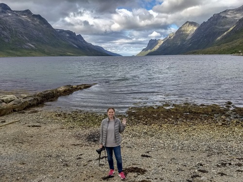 Sarah, a woman, standing in front of a gorgeous Fjord (water and mountains) in Norway. She's also holding a camera.